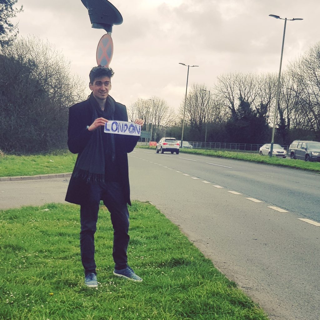 Young man hitchhiking on the side of a road holding a handmade sign that says 'London'