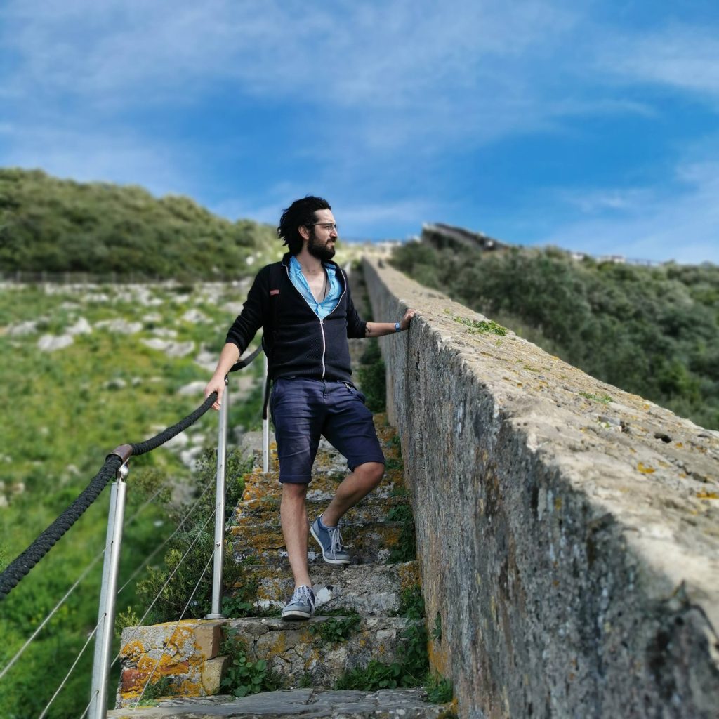 Man walking up ancient stone steps beside a high wall, surrounded by greenery under a blue sky.