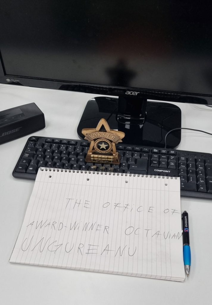 Computer desk with handwritten sign saying “The office of award-winner Octavian Ungureanu,” winner trophy, keyboard, and monitor.