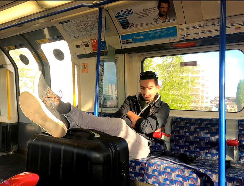 Man sitting on the London Underground with legs up on a suitcase, looking out the window on a sunny day.
