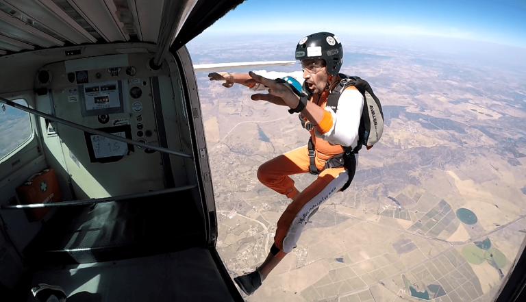 A skydiver in an orange jumpsuit taking the leap from the plane door, a look of focused intensity on his face as he overcomes the fear of jumping.