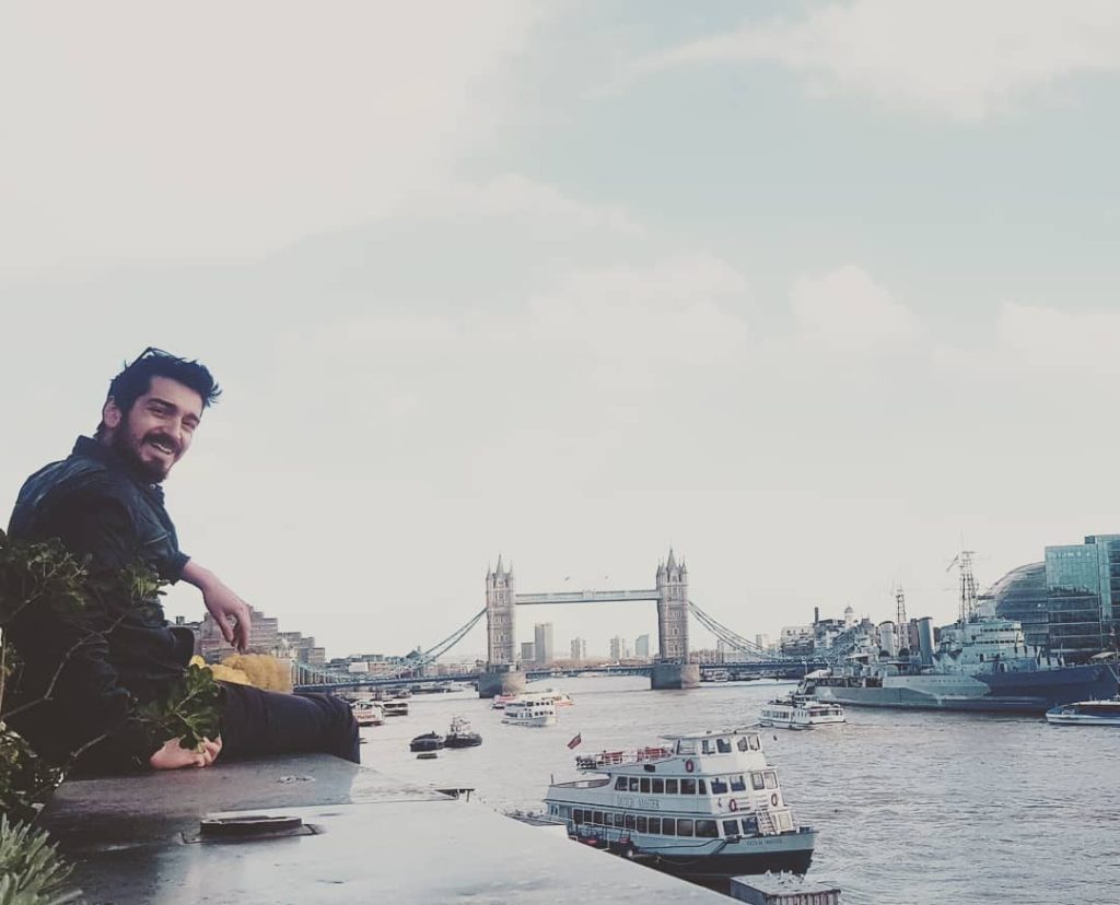 A man sitting by the River Thames in London, looking back at the camera with Tower Bridge in the background. This represents the 'before' stage of a journey with post achievement depression.